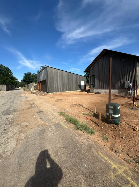 Rural dirt road beside dark metal sheds under a blue sky with clouds