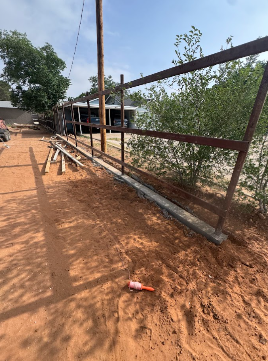 Rusty metal gate frame beside a dirt road with scattered boards and trees in the background