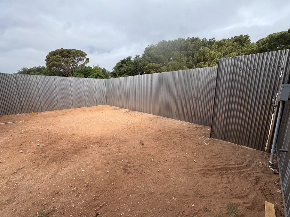 Empty dirt yard enclosed by gray corrugated metal fencing under a cloudy sky