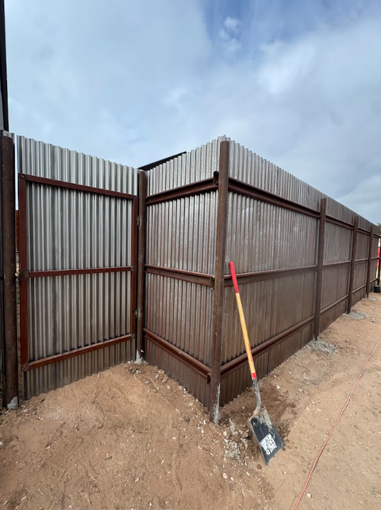 Corrugated metal fence corner beside dirt ground, with a shovel leaning against it under a cloudy sky