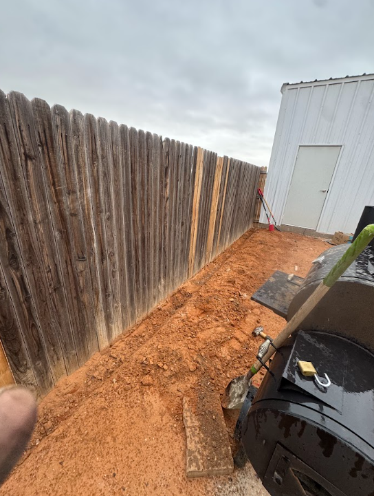Narrow dirt path beside a wooden fence and white shed, with a grill in the foreground.