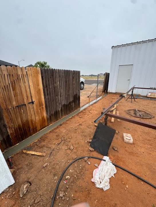 Outdoor dirt yard beside a white building, with a wooden fence, hose, and scattered construction debris.