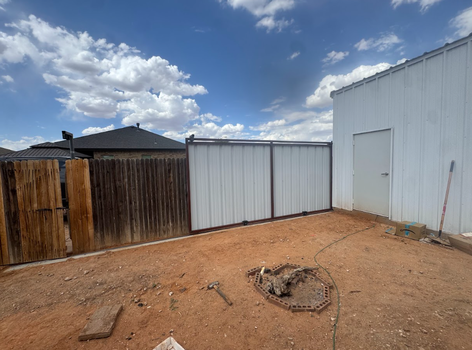 Small backyard with dirt ground, corrugated metal fence, white shed, and blue sky with clouds.