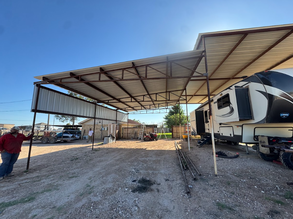 RV parked under a metal carport in a gravel lot, with a person standing at left and a clear blue sky.
