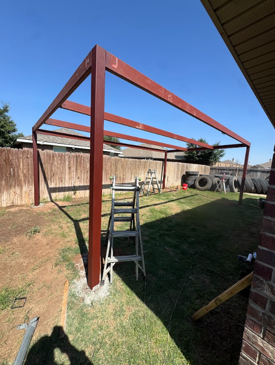 Backyard steel frame structure being built, with a ladder beneath it and a fence in the background