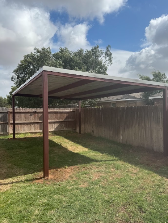 Covered backyard patio with a metal roof, wooden fence, grass, and cloudy sky.