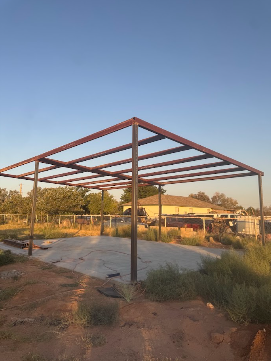 Partially built metal frame on a concrete pad at sunset, with dry grass and trees nearby