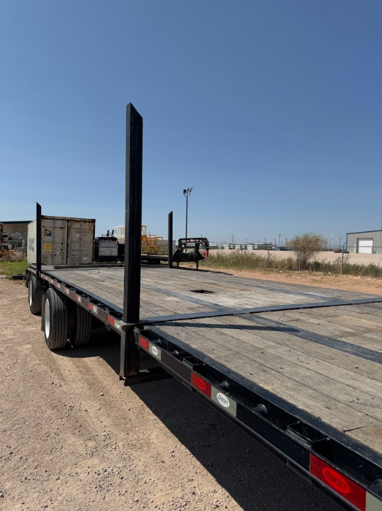 Black flatbed trailer parked on a gravel lot under a clear blue sky.