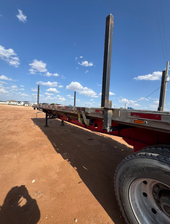 Flatbed trailer parked on a dirt lot under a blue sky with scattered clouds.
