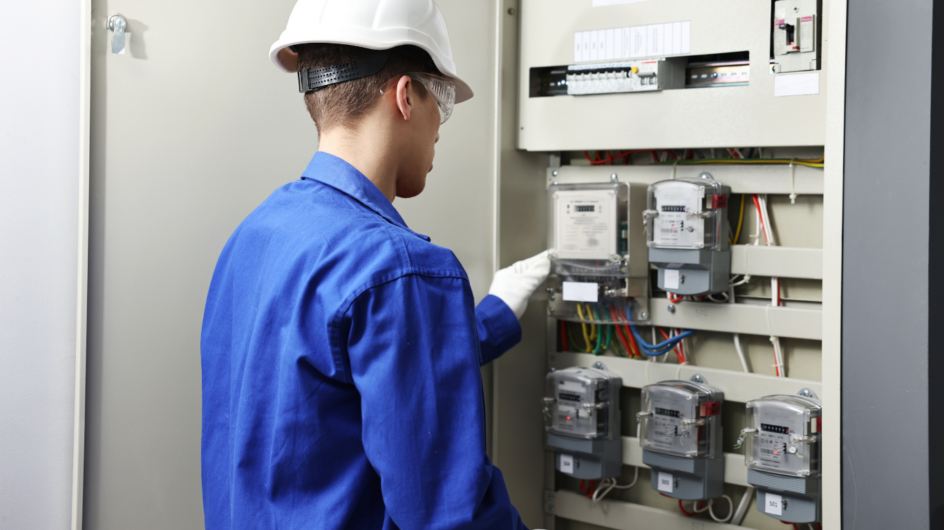 Technician in blue coveralls and hard hat inspecting an electrical control panel with meters and wiring