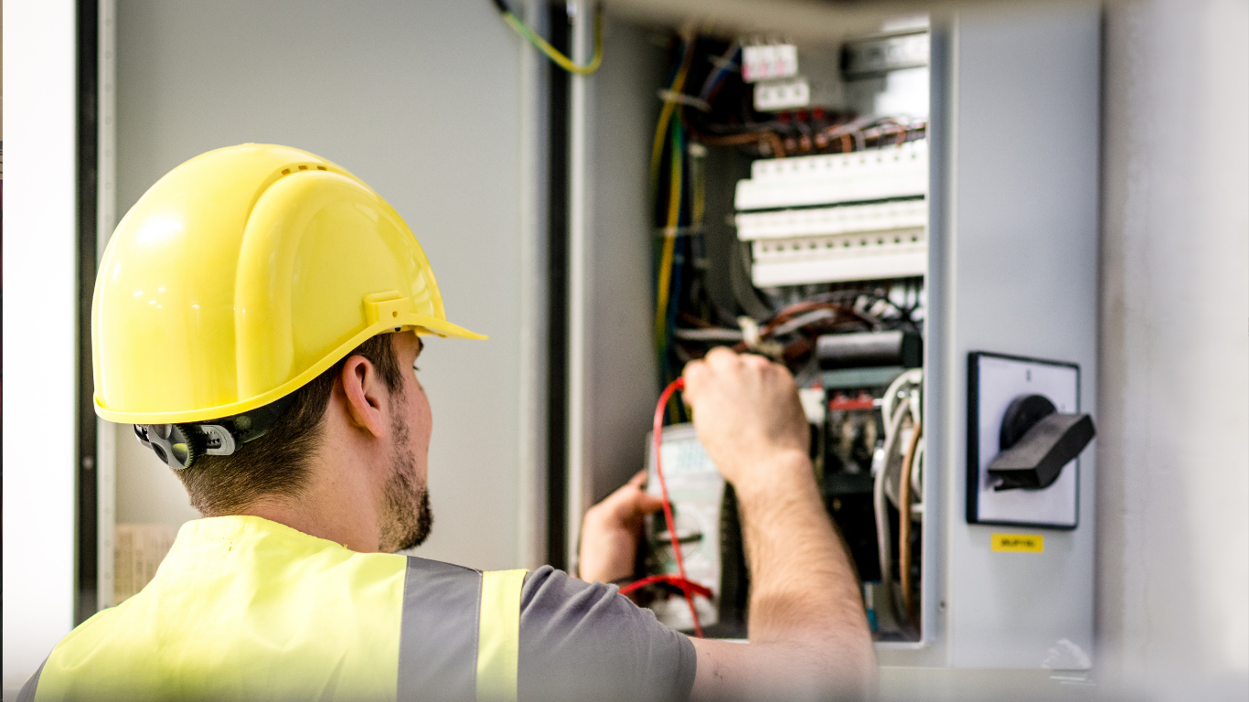 Electrician in yellow hard hat inspecting wiring inside an open electrical panel
