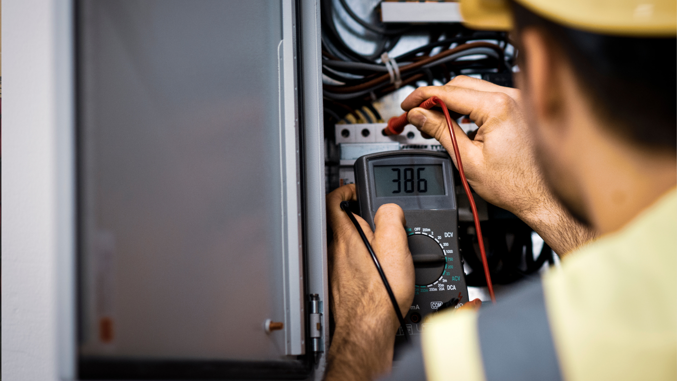 Technician inspecting electrical wiring in a control panel, using a multimeter inside a gray cabinet.