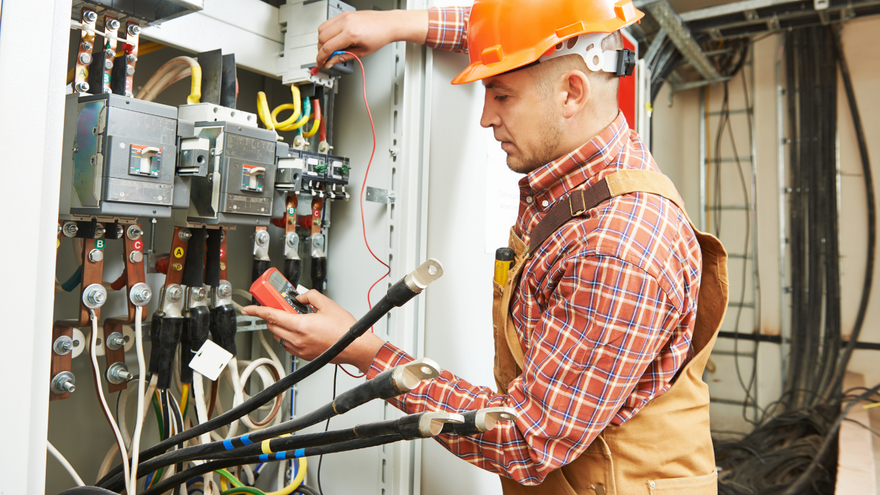Electrician in orange hard hat inspecting control panel and wiring in an industrial facility