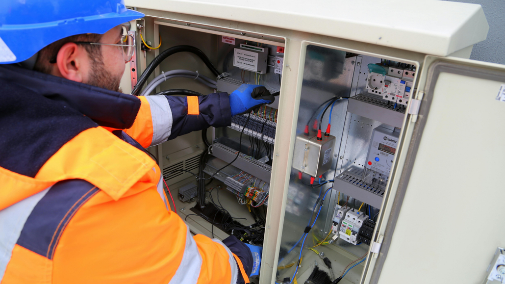 Technician in orange jacket working inside an open outdoor electrical control cabinet