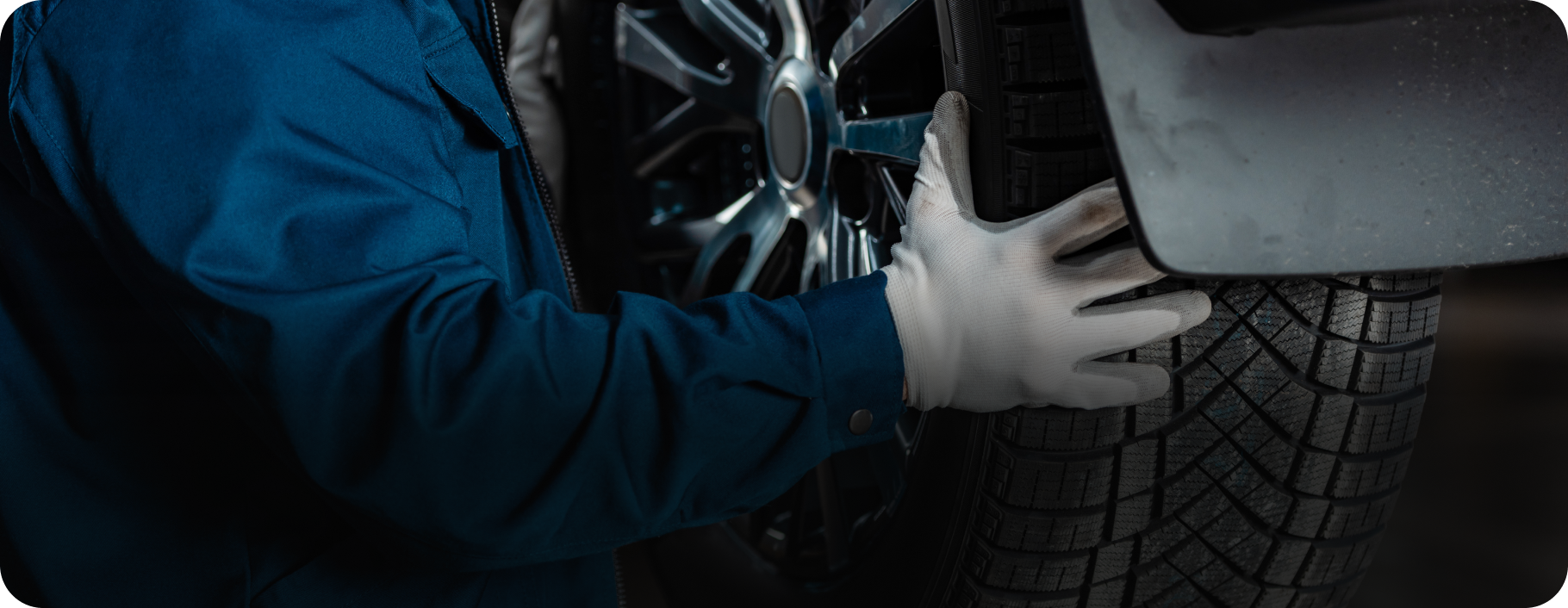 Mechanic in blue coveralls and white gloves checks a tire near the car's bumper.