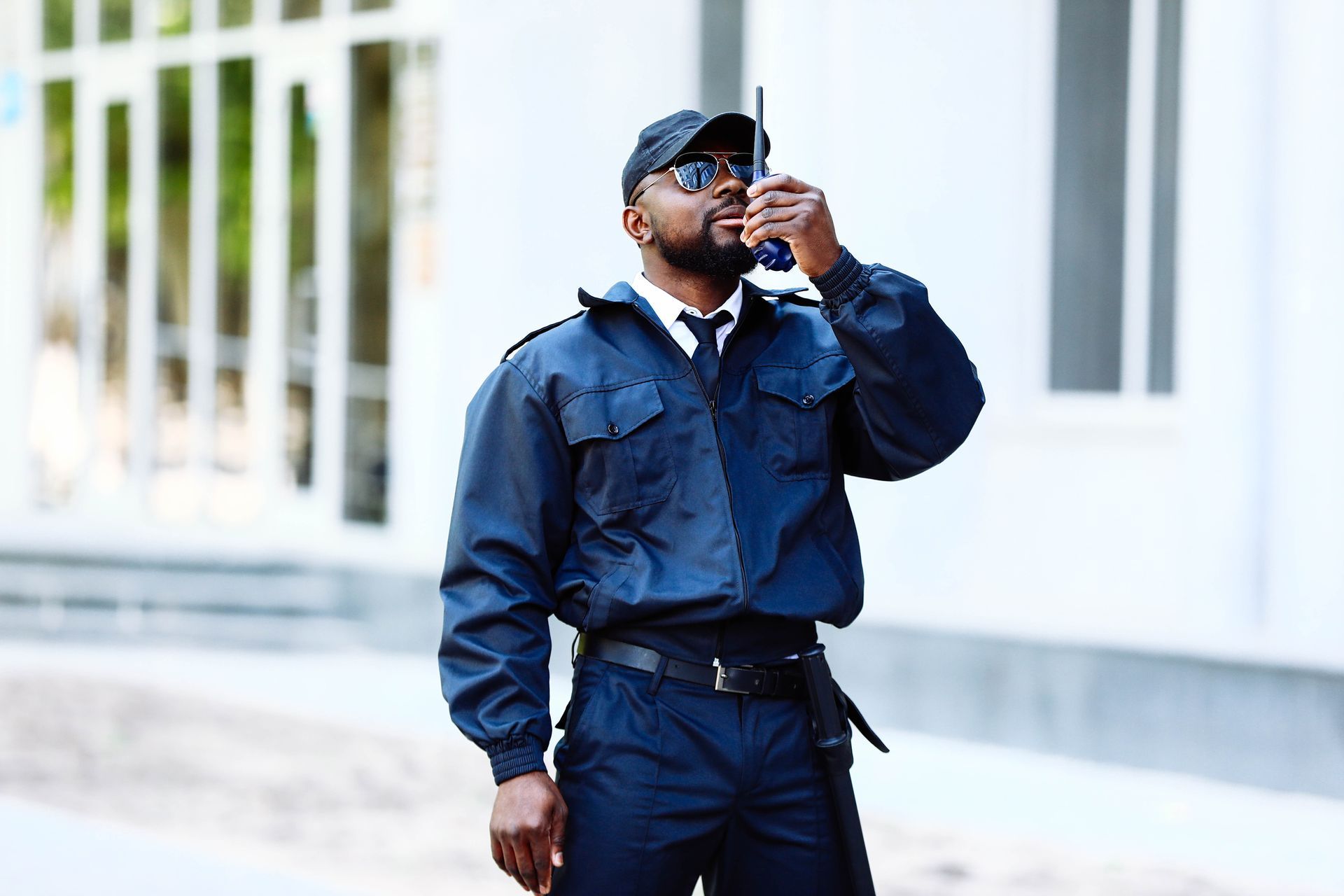 Uniformed security worker holding a handheld radio while standing outdoors near a building.