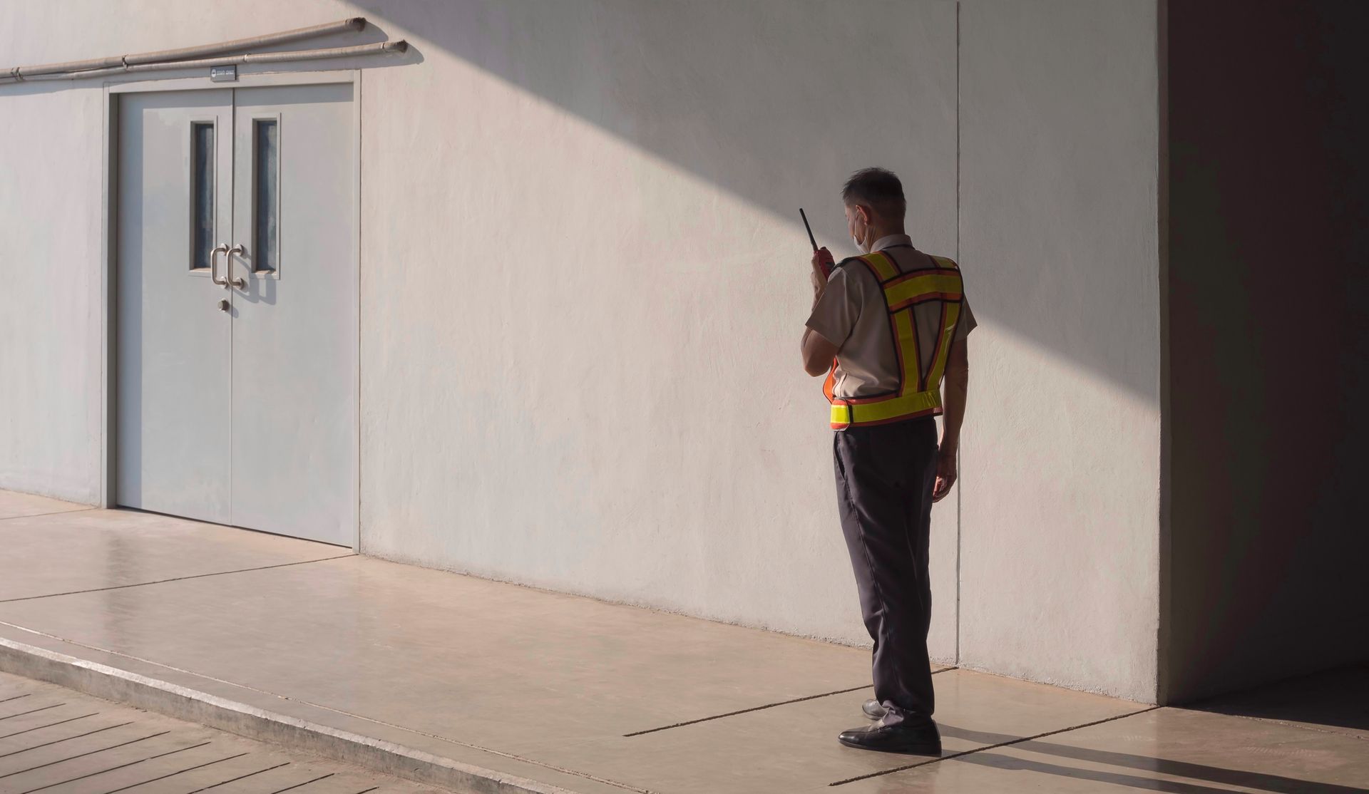 Uniformed worker wearing a safety vest uses a handheld radio while standing near a building. Uniformed worker wearing a safety vest uses a handheld radio while standing near a building.
