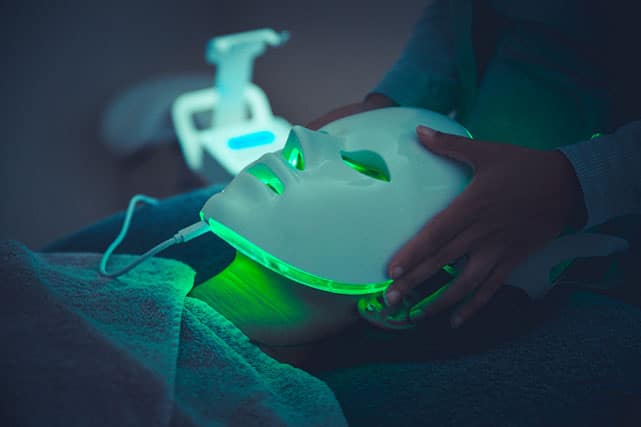 Gloved hands holding a glowing green facial LED mask in a dark spa setting
