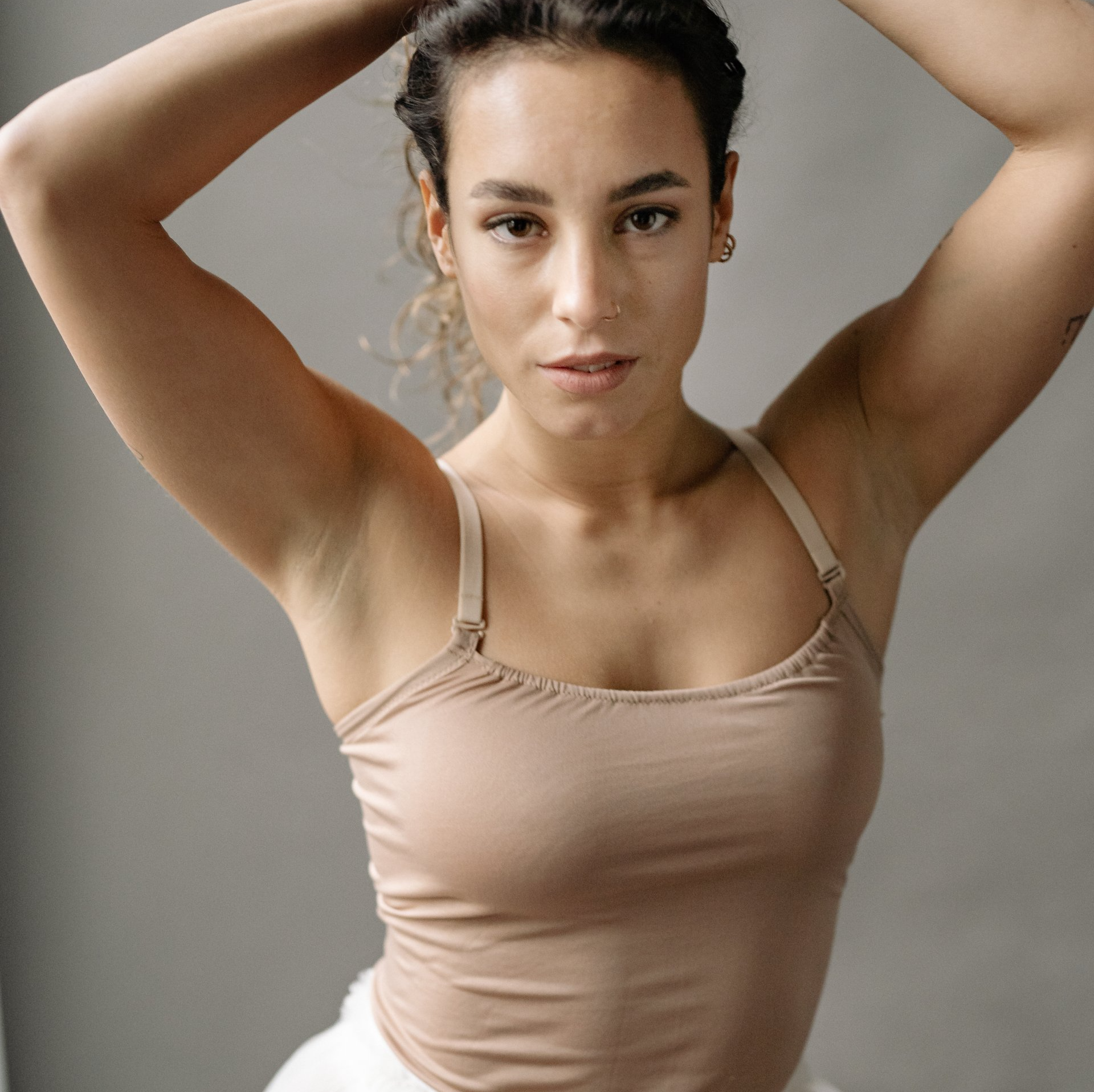 Woman posing with arms raised, wearing a beige tank top against a plain gray background