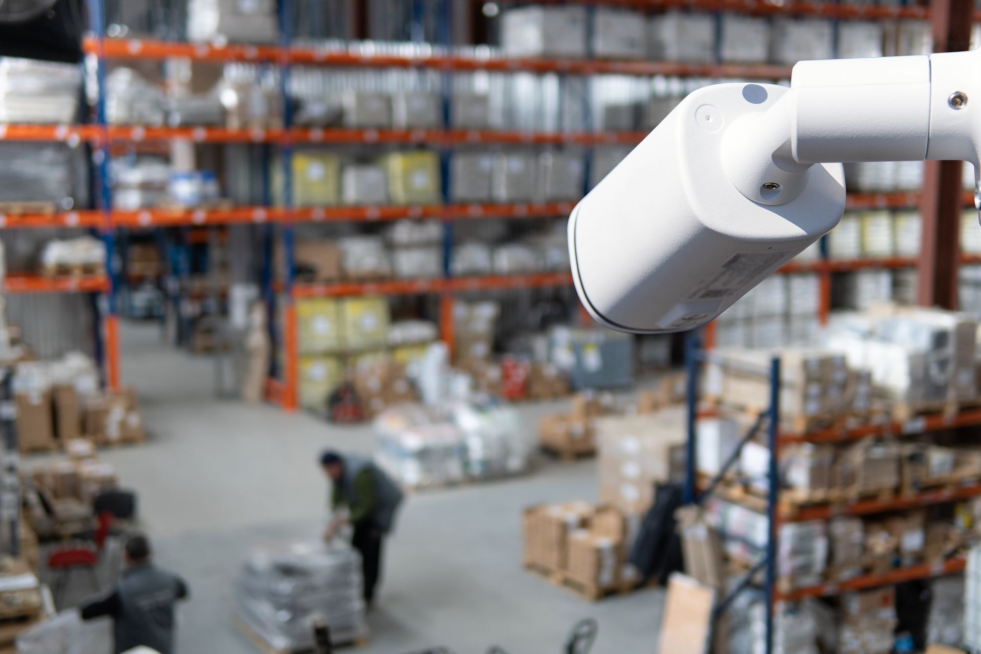 Warehouse aisle with stacked boxes and shelves, viewed past a large white robotic arm in the foreground
