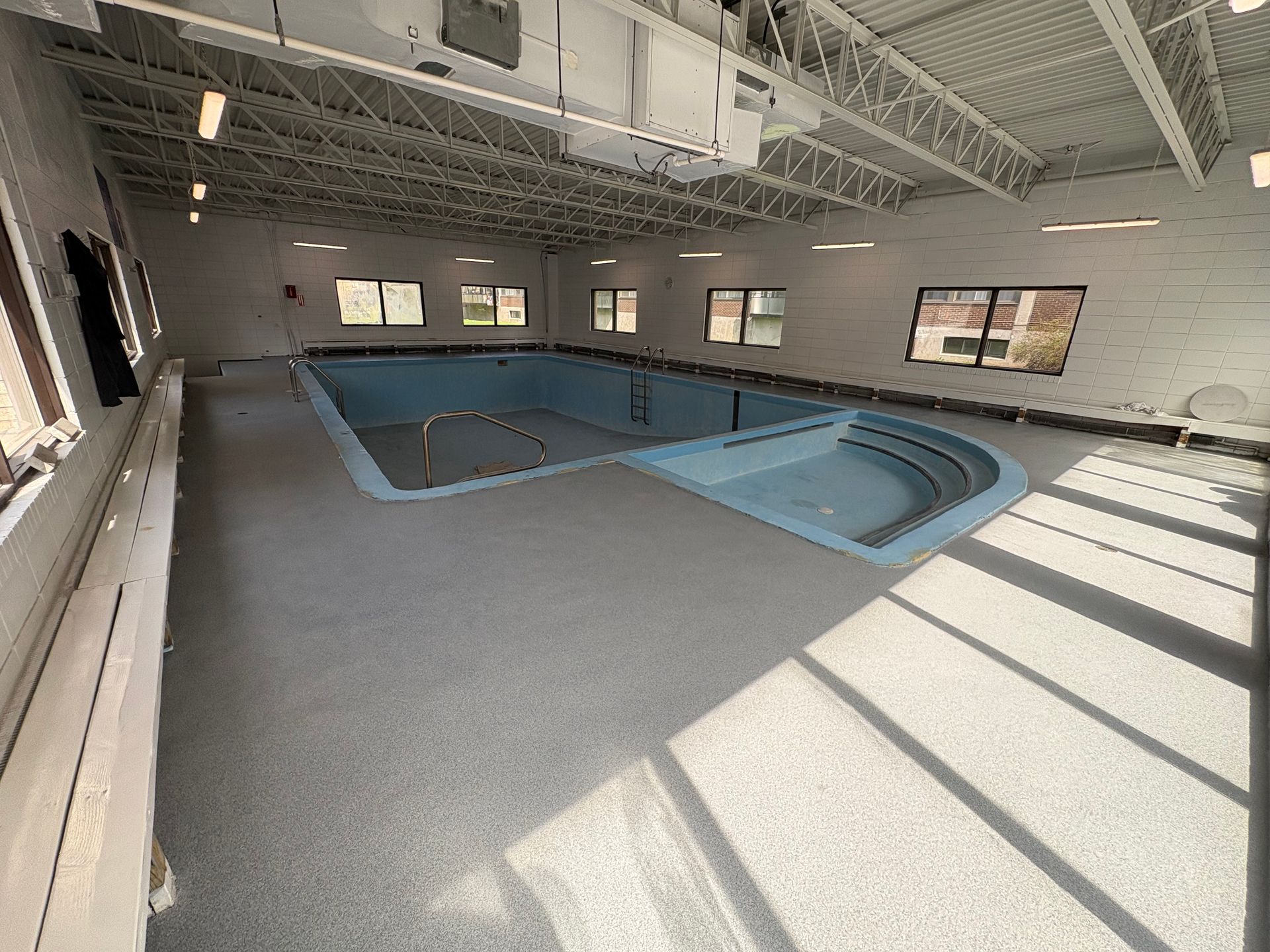 Empty indoor swimming pool in a white-walled room with gray flooring and windows.