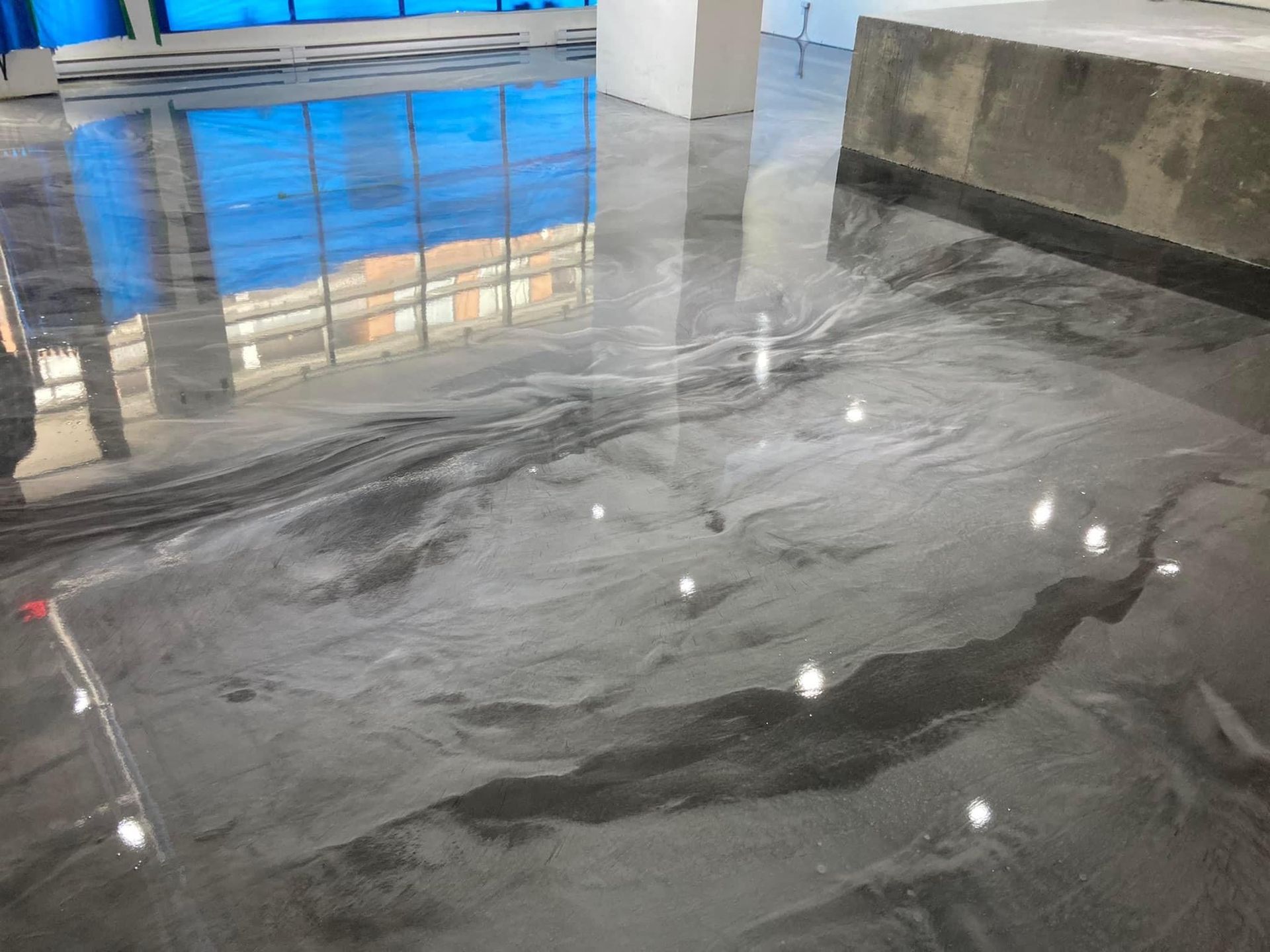 Sol époxy gris métallisé brillant avec des motifs fluides, reflétant des fenêtres bleues et un pilier blanc dans un bâtiment.