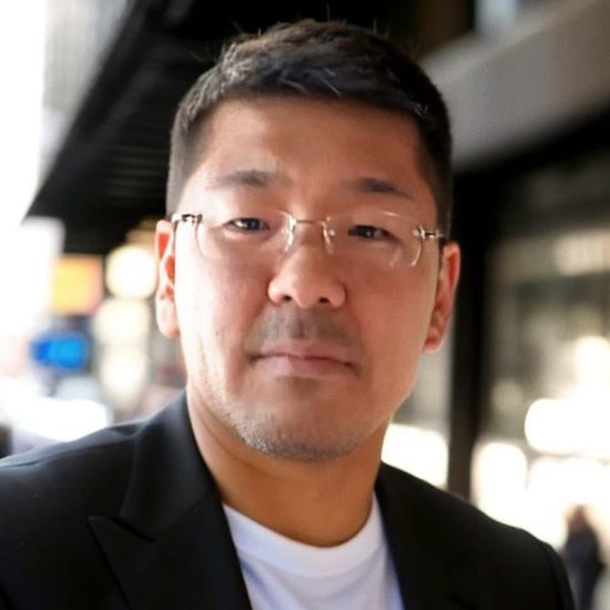 Close-up portrait of a man wearing glasses and a dark blazer in an outdoor setting.