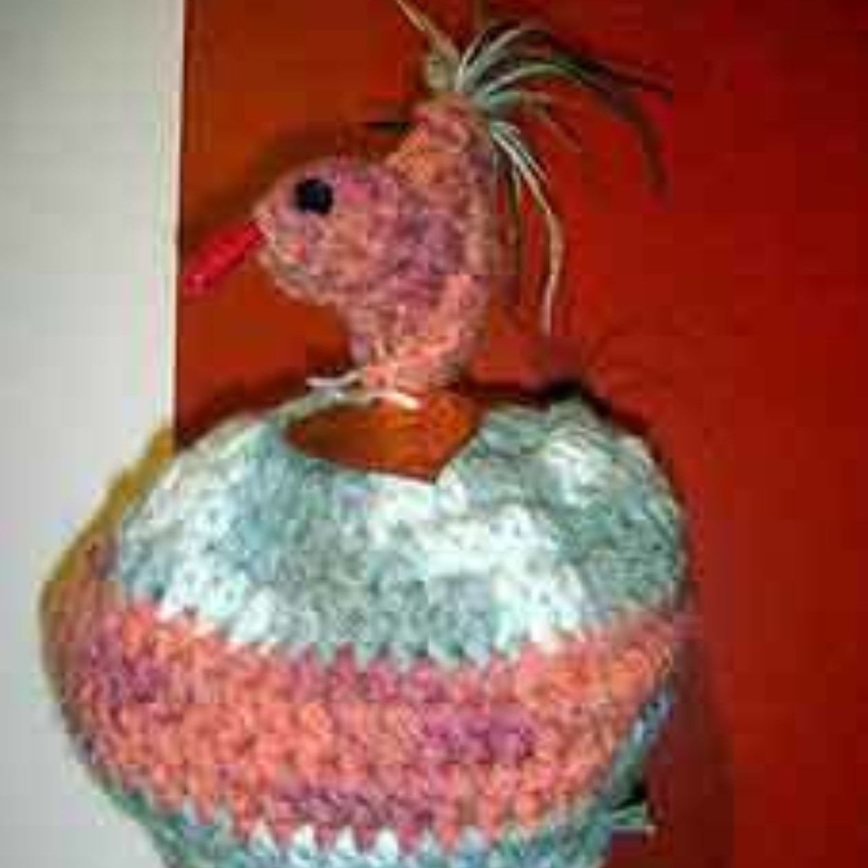 A crocheted bird with a red beak is hanging on a red wall.