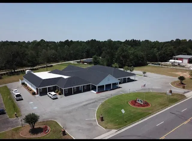 Aerial view of a single-story commercial building with a blue facade, black roof, and manicured landscaping, set against a backdrop of trees.