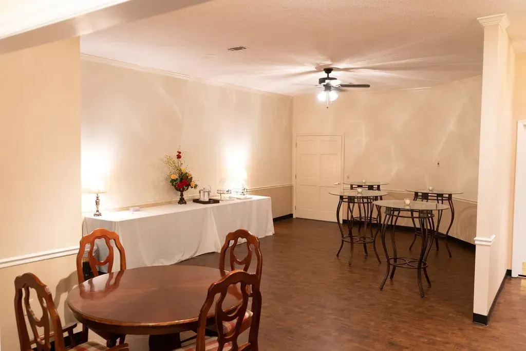 Empty event space with tables, chairs, and a buffet table. Cream-colored walls, brown floor, ceiling fan.