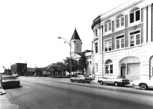 Street view with parked cars, buildings, and a clock tower in the distance.