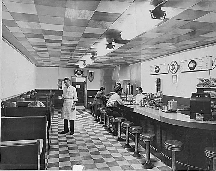 Diner interior with checkerboard floor and ceiling, booths, counter, customers, and a server in white apron.