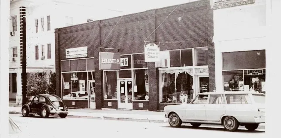 Black and white photo of a row of storefronts on a street, with parked cars and a Beetle.
