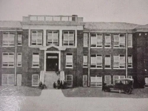 Brick building with multiple windows; people near entrance, vintage car parked nearby.