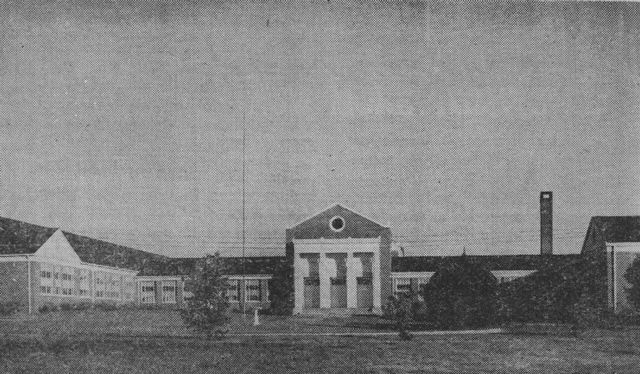 Exterior view of a large, two-story brick building with a central portico and tall smokestack.