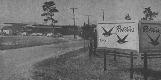 Two signs for Robbins Packing Co. with swallows, near a building and road, black and white photo.