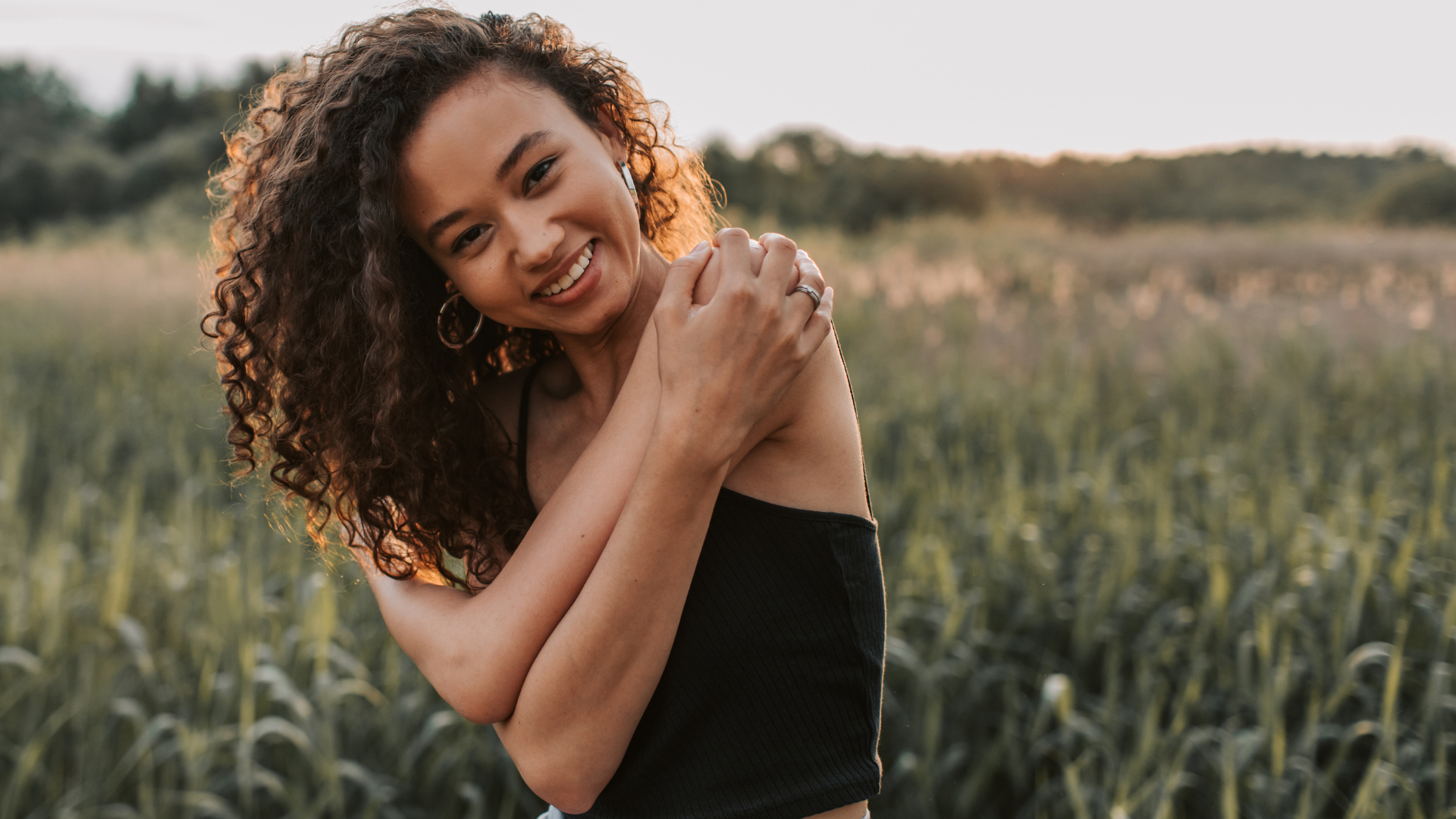 Smiling woman in a black top standing in a field at sunset, holding one shoulder