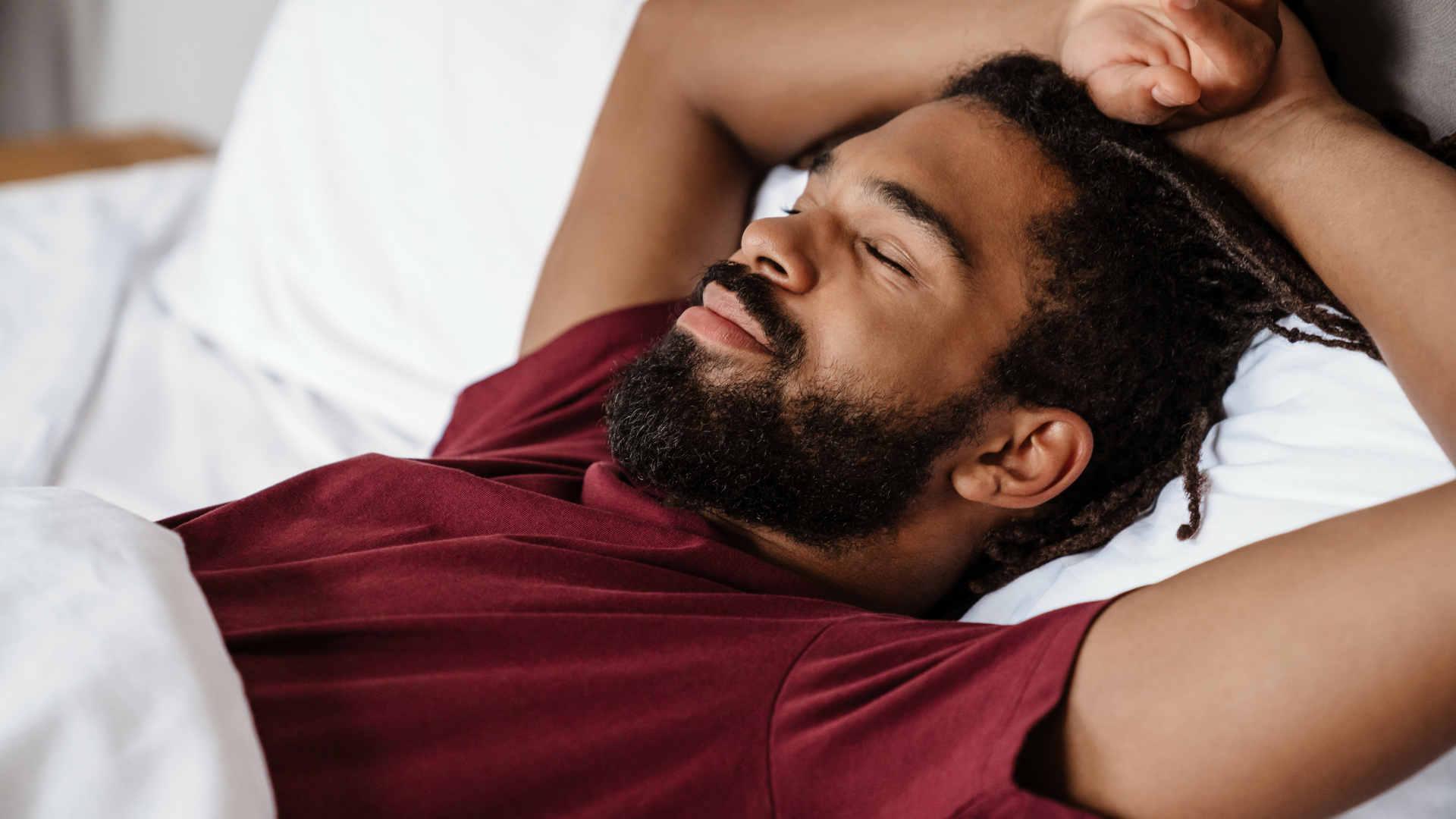 Man sleeping on a white bed with arms behind his head, wearing a maroon shirt