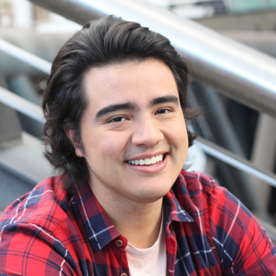 Smiling person in a red plaid shirt seated indoors against a blurred railing background