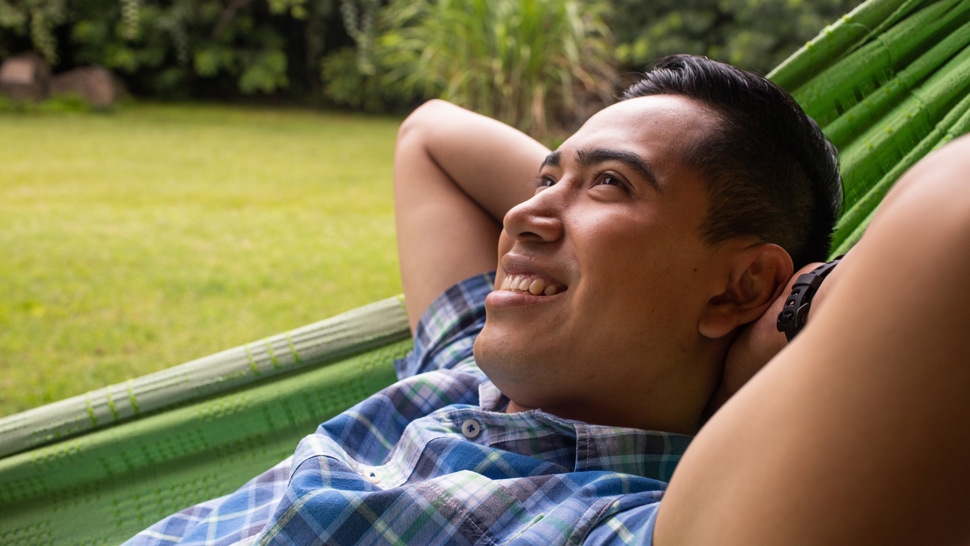 Man relaxing in a green hammock outdoors, smiling with hands behind his head.
