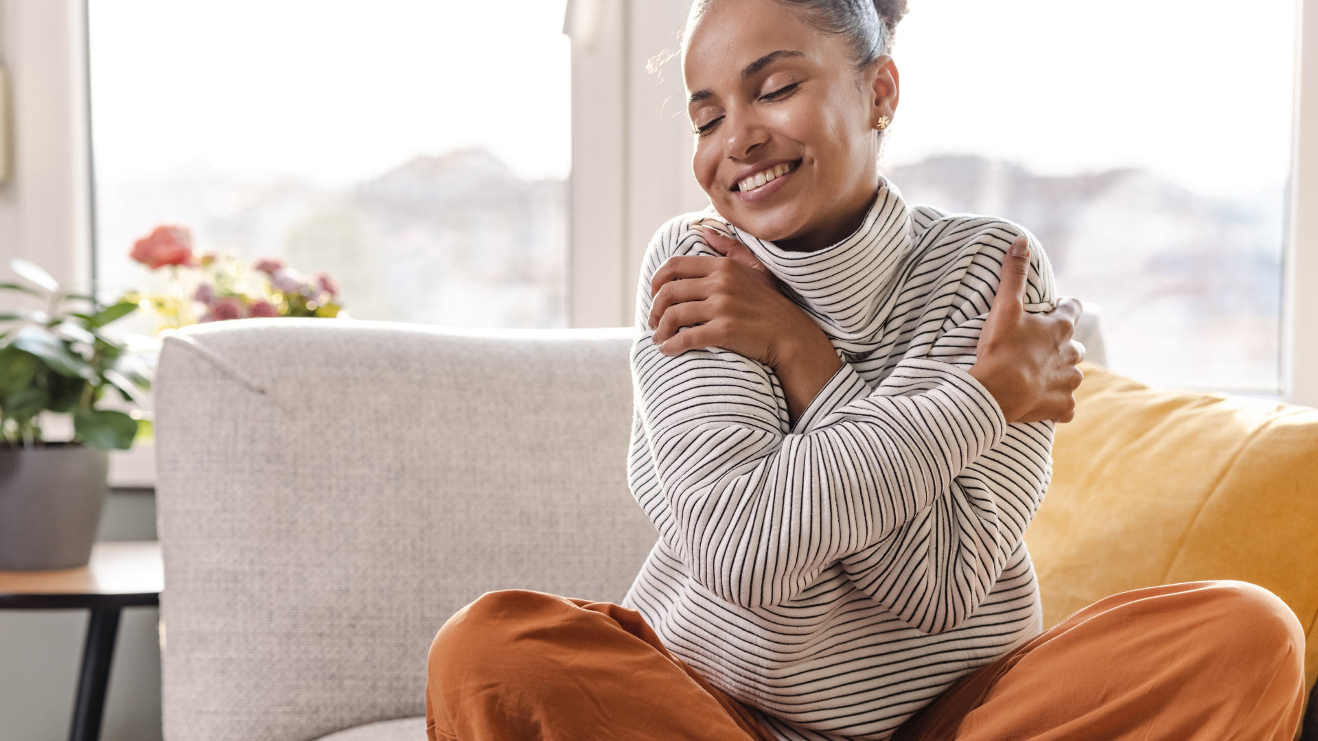 Smiling person in a patterned sweater hugs themselves while sitting on a sofa in a bright living room