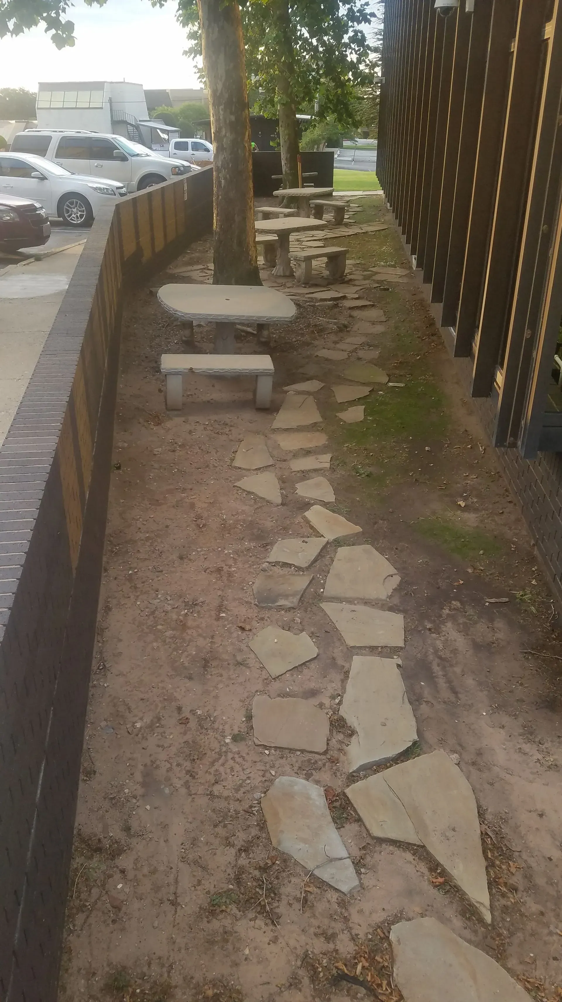 Stone path between a brick wall and outdoor seating area with trees. Cars visible in background.