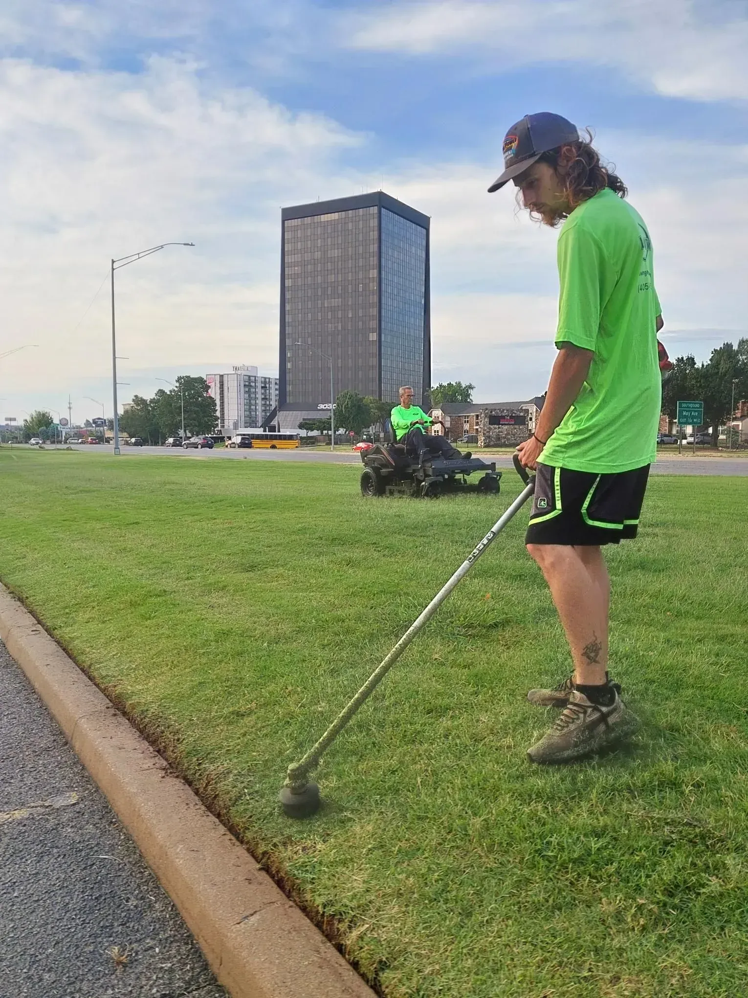 A person in green shirt using a weed whacker on a grassy median next to a road, skyscraper in background.