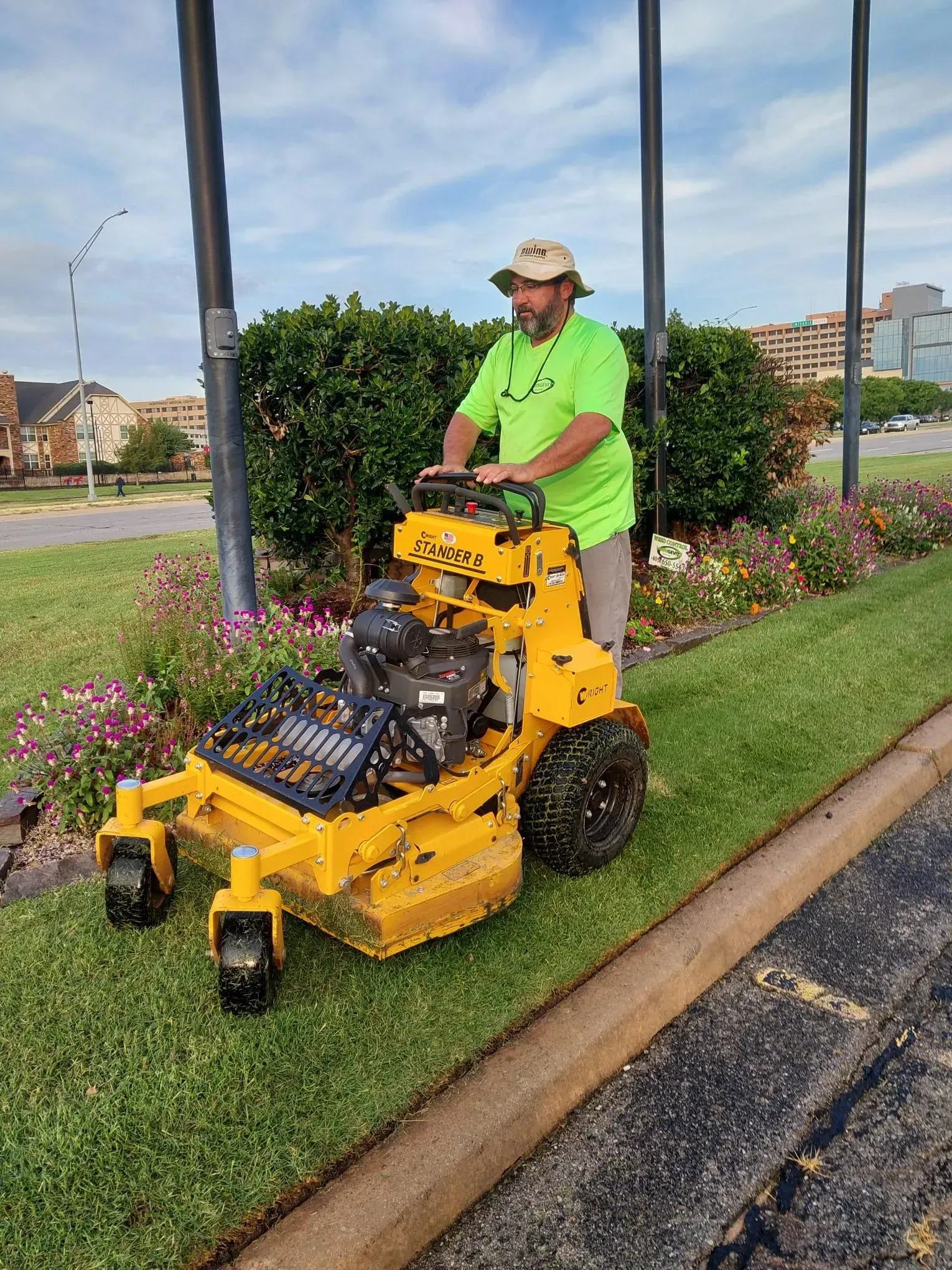 Man operating yellow lawnmower, cutting grass next to a curb. Green shirt, beige hat, and flowers in the background.