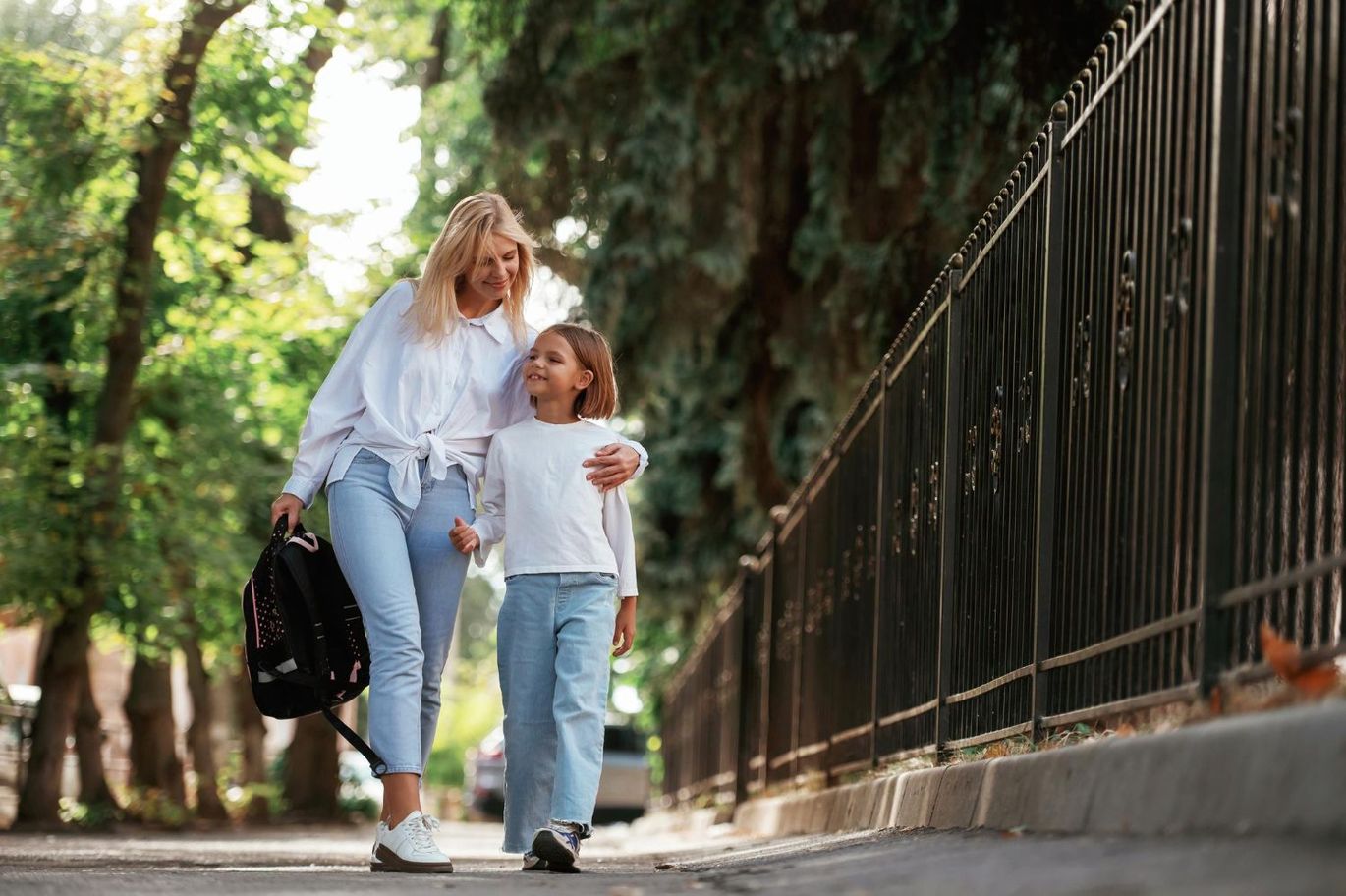 Woman and child walking on a sunny sidewalk by a black iron fence, holding hands and carrying bags.