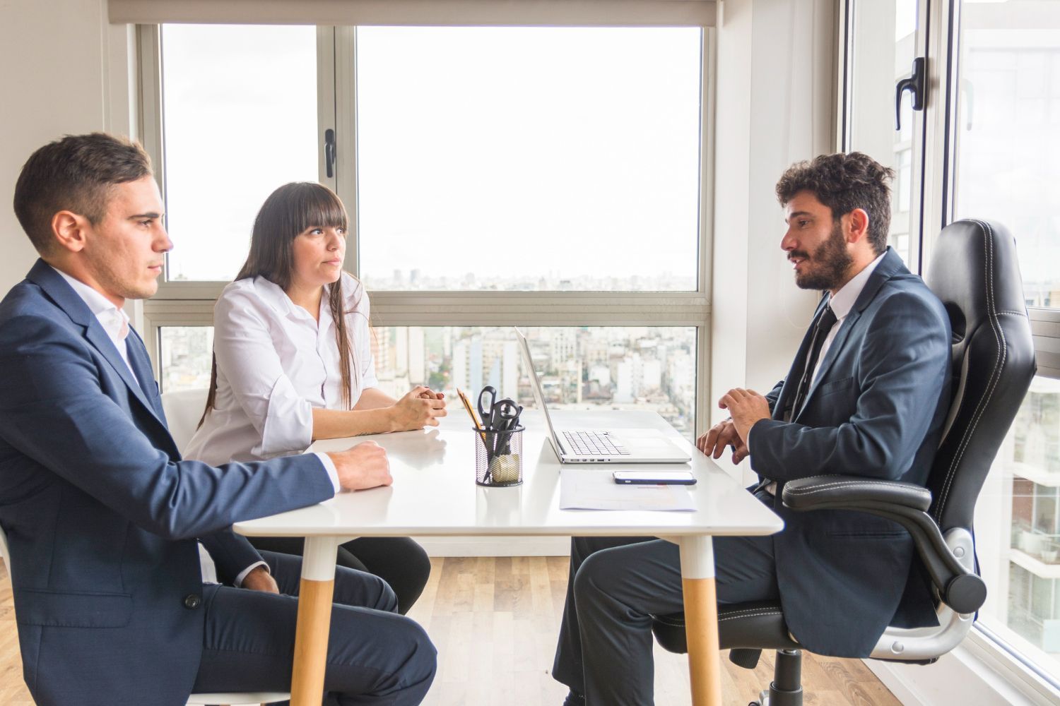 Three people in a bright office meeting across a white table with a laptop and notebook