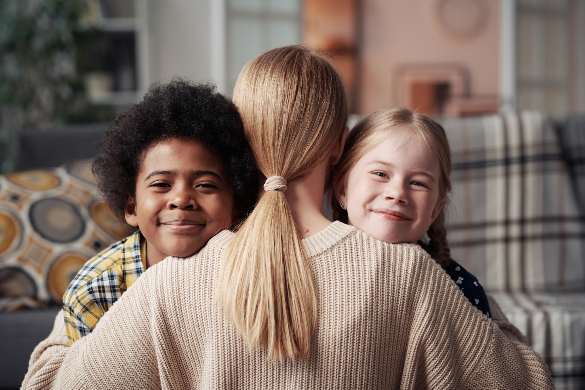Two smiling children hugging a woman from behind on a couch in a cozy living room