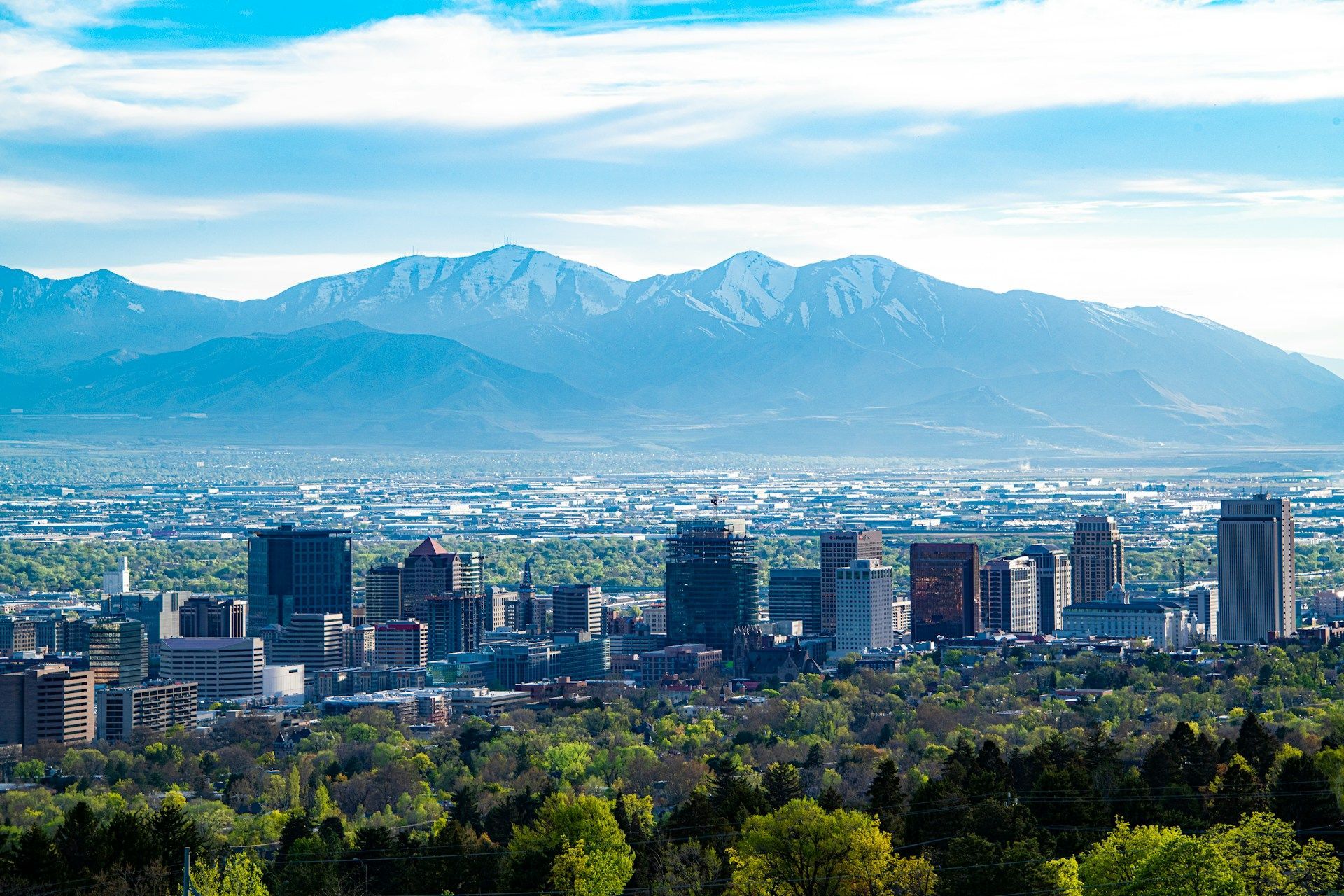 City skyline with green trees in front and snow-capped mountains in the distance under a clear blue sky