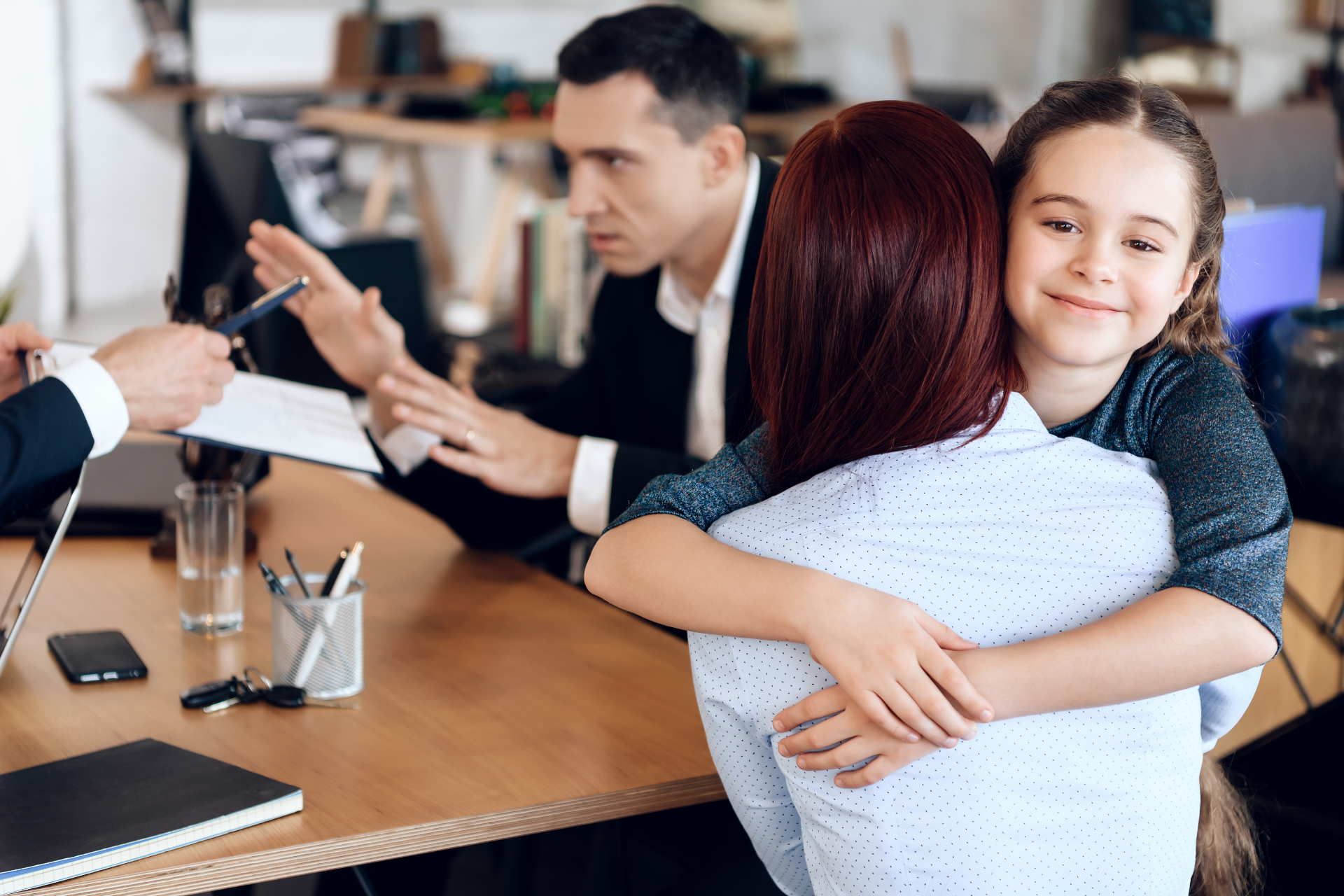 Woman hugging a smiling child in an office while two people discuss paperwork at a table