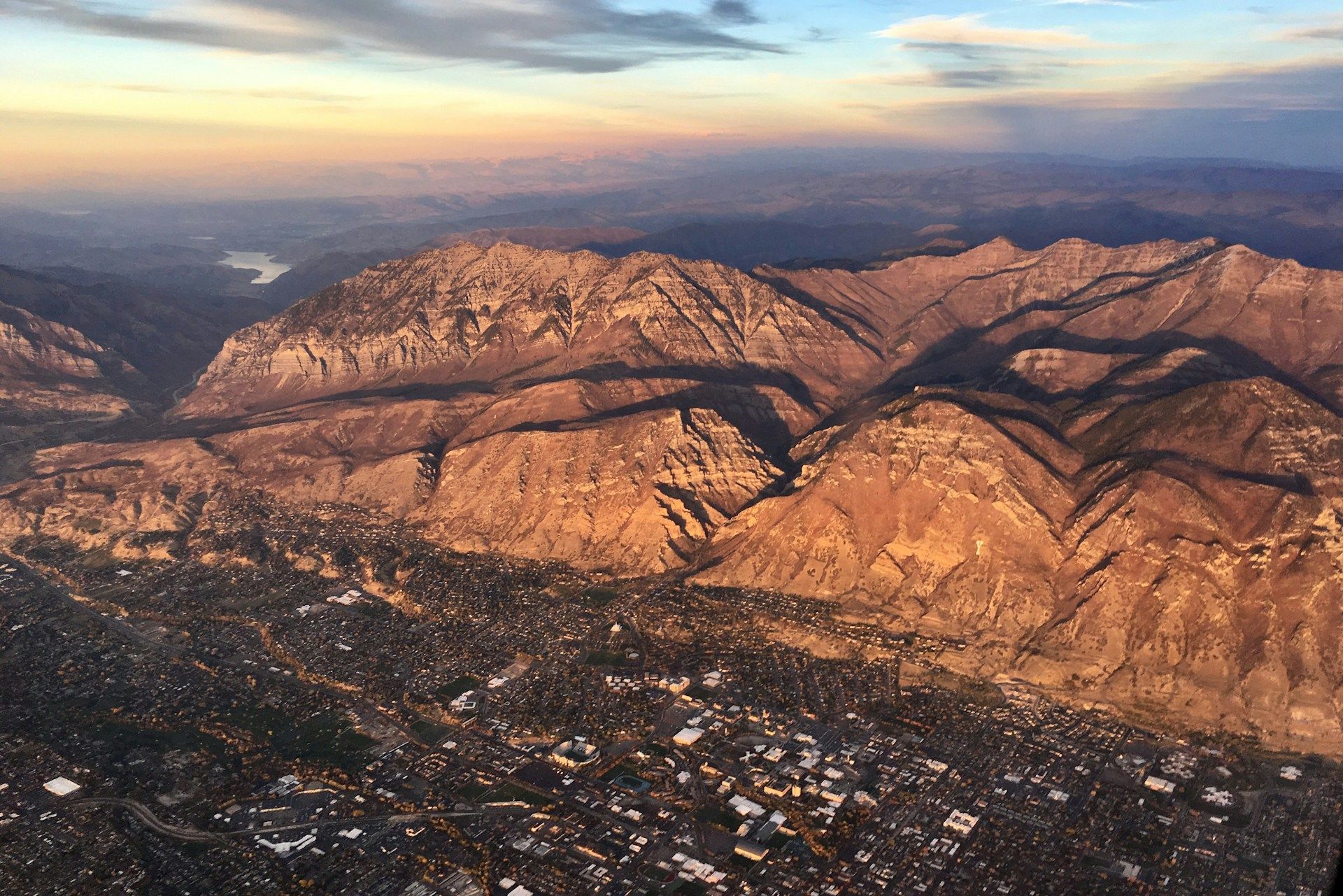 Sunlit rocky mountains overlooking a town at dusk, with a distant valley and cloudy sky